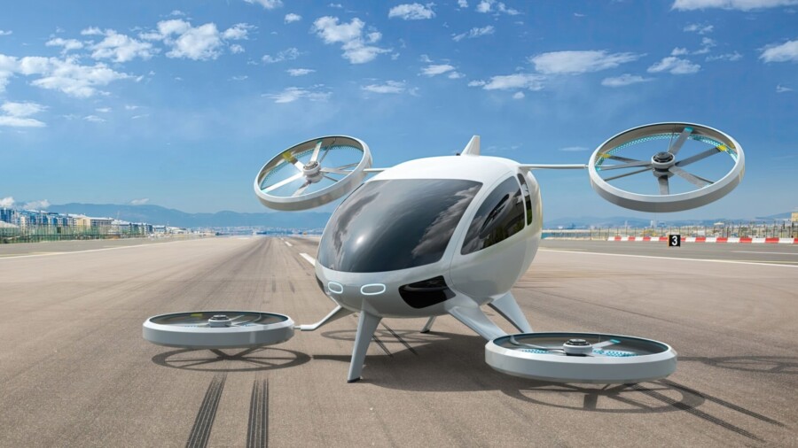 A white electric powered Vertical Take Off and Landing eVTOL aircraft with four rotors parked on the empty runway of an airport, with distant buildings visible in the background.