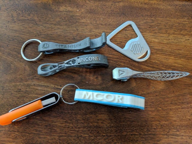 [3D printed bottle openers circa 2018, with models from 3D Systems (printed in titanium and Inconel), Markforged, Velo3D, and – 3D printed in paper with an attached media kit USB – MCor // Image Source: Author]